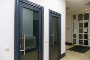 A split-screen comparison showing an anodized aluminum door on the left with a metallic, dark bronze-black sheen and visible grain, next to a powder-coated aluminum door on the right with a solid, uniform matte black finish, both installed in exterior masonry walls.